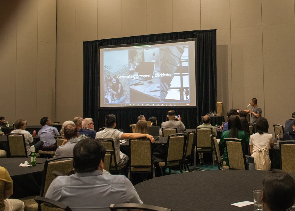 A dinner hall of round tables facing a large screen showing a presentation slide